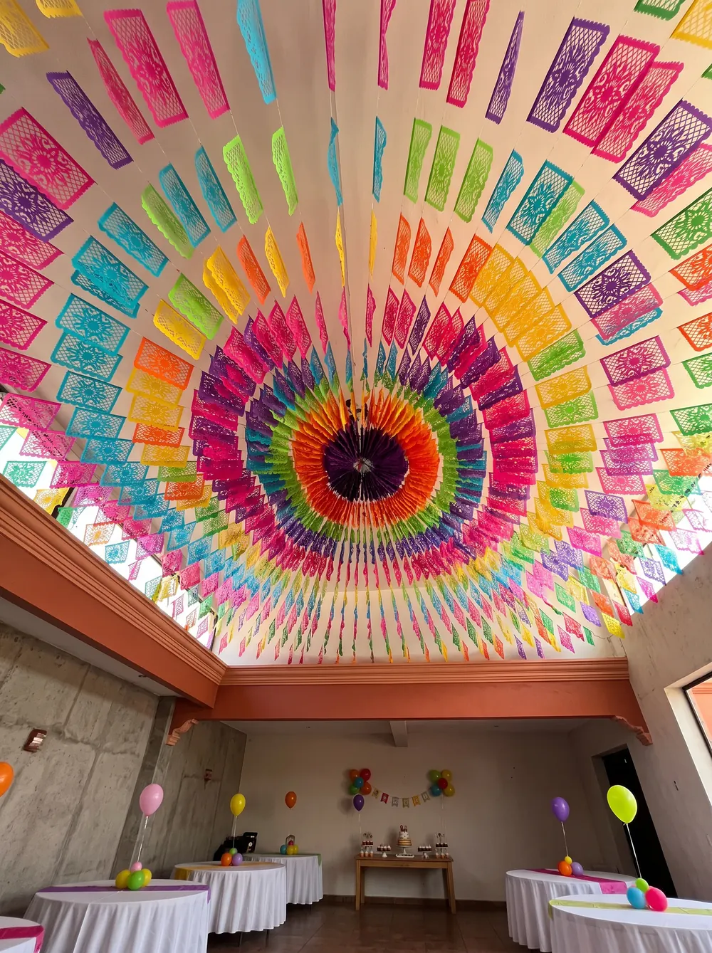 Salón de fiesta infantil con el techo cubierto por un arreglo circular de papel picado multicolor que sale del centro hacia las cuatro paredes; abajo, mesas redondas con mantel blanco y globos